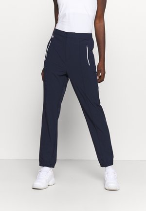 Personne portant un pantalon bleu marine avec des poches zippées blanches et des baskets blanches, debout devant un fond clair uni.