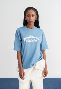 Camiseta de algodão azul com um logotipo branco "New Balance Athletics", mangas curtas e um corte solto, combinada com calças na cor creme.