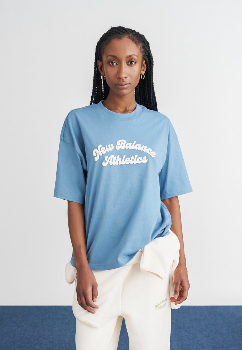 Camiseta de algodão azul com um logotipo branco "New Balance Athletics", mangas curtas e um corte solto, combinada com calças na cor creme.