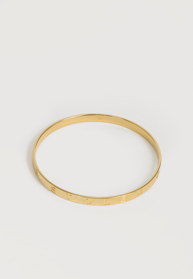 Χρυσό βραχιόλι με εγχάρακτα γράμματα που σχηματίζουν τη λέξη «STOLEN» σε απλό ανοιχτό φόντο.