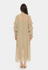 Beige, lätt, genomskinlig lång klänning med lager av volanger på ärmarna och knytning bak. Mjuk textur med en flödande form.