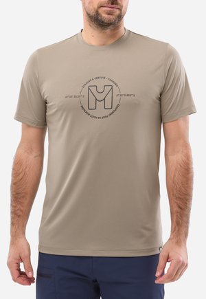 Homme portant un t-shirt beige à manches courtes avec un logo circulaire et du texte en français, associé à un pantalon bleu marine, debout devant un fond blanc.