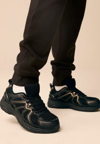 Baskets de sport noires en mesh et en matériau synthétique, avec des accents dorés sur les côtés, et une semelle texturée. Associées à des joggings sombres.