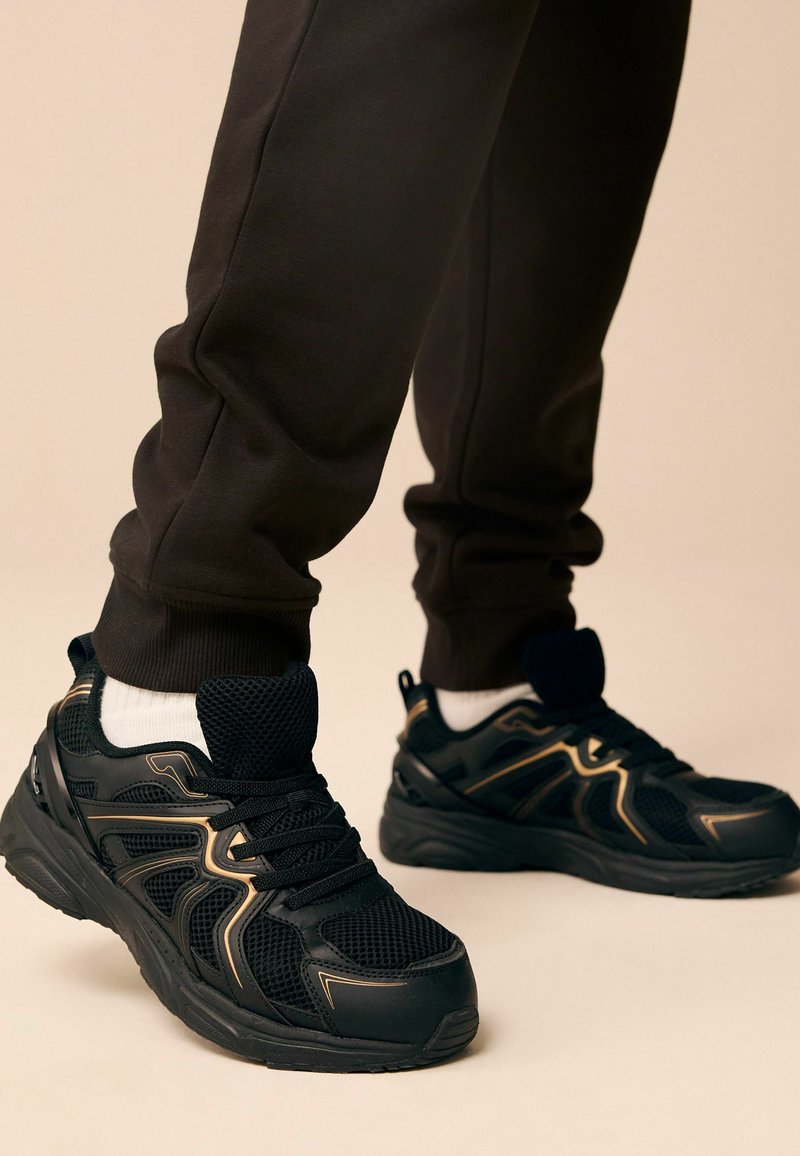 Baskets de sport noires en mesh et en matériau synthétique, avec des accents dorés sur les côtés, et une semelle texturée. Associées à des joggings sombres.