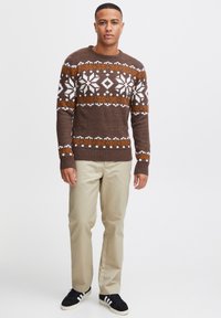 Suéter marrón de punto con patrones de copos de nieve en blanco y naranja, cuello redondo y puños acanalados. Combinado con pantalones beige claros y zapatos negros.