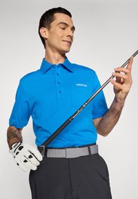 Blå polo skjorte med knappet krage og lomme, kombinert med grå belte og svarte bukser. Holder en golfkølle; bruker en hvit golfhanske.