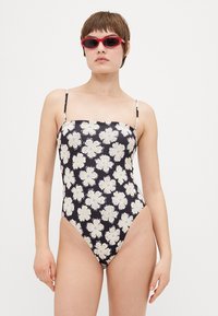 Einteiliger Badeanzug mit dünnen Trägern, schwarzem Stoff mit großen weißen Blumenmustern, hohen Beinausschnitten und einer engen Silhouette.