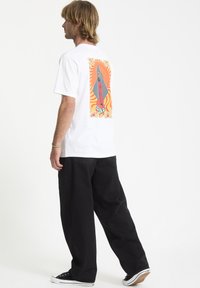 Hvid kortærmet t-shirt med et farverigt grafisk print på ryggen, der har fede linjer og svirlende mønstre. Bæres med løse sorte bukser.