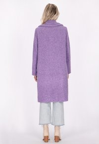 Cappotto viola peloso con maniche lunghe e colletto, che arriva fino a metà coscia. Indossato sopra jeans chiari strappati e stivali con tacco beige.