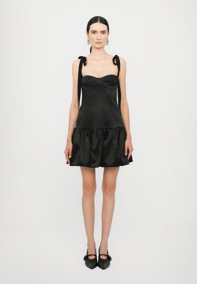 Vestido de satén negro con un corsé ajustado, dobladillo con volantes y lazos en los hombros. Los zapatos son negros de punta con un detalle de lazo.