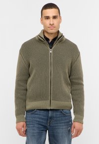 Olivgrön zip-up tröja med vertikal ribbstruktur, hög krage och åtsittande muddar, matchad med blå slitna jeans.