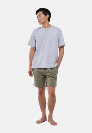 Homme debout pieds nus portant un t-shirt gris clair et un short vert olive avec une main dans la poche, regardant sur le côté.