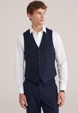 Muž v tmavomodrej formálnej veste na bielom košeľovom tričku a tmavomodrých nohaviciach, pozerajúci sa do strany na jednoduchom pozadí.