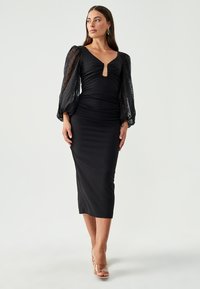 Black midi dress with a fitted silhouette, deep V-neck, and ruching. Sheer, puffed sleeves with polka-dot texture. Clear strappy heels.