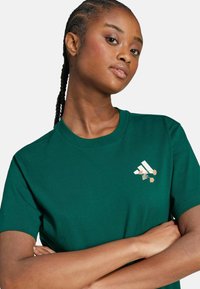 Camiseta de algodón verde con cuello redondo, que presenta un logo de rayas blancas y bordados florales en el pecho. Mangas cortas y corte relajado.