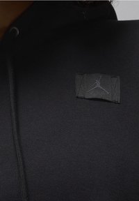 Sweat à capuche noir en tissu texturé, avec un petit écusson du logo Jordan noir sur la poitrine. Comprend une fermeture éclair avant et une capuche à cordon.