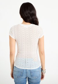 Mujer con cabello largo y oscuro de espaldas, vistiendo una blusa blanca de encaje de manga corta y vaqueros azul claro con pulseras en la muñeca izquierda.