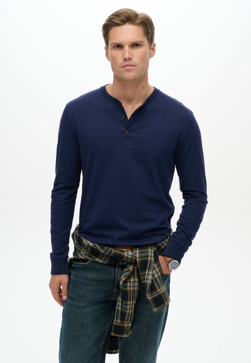 Young man wearing a navy long-sleeve henley shirt with a plaid shirt tied around his waist and blue jeans, standing against white background.
