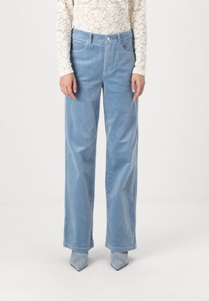 Person wearing light blue wide-leg corduroy pants, white lace long-sleeve top, and pointed-toe light blue shoes, standing against plain background.