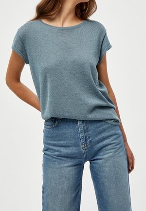 Frau trägt ein hellblaues Kurzarm-Stricktop, das in hoch taillierte blaue Jeans gesteckt ist, vor einem einfarbigen Hintergrund.