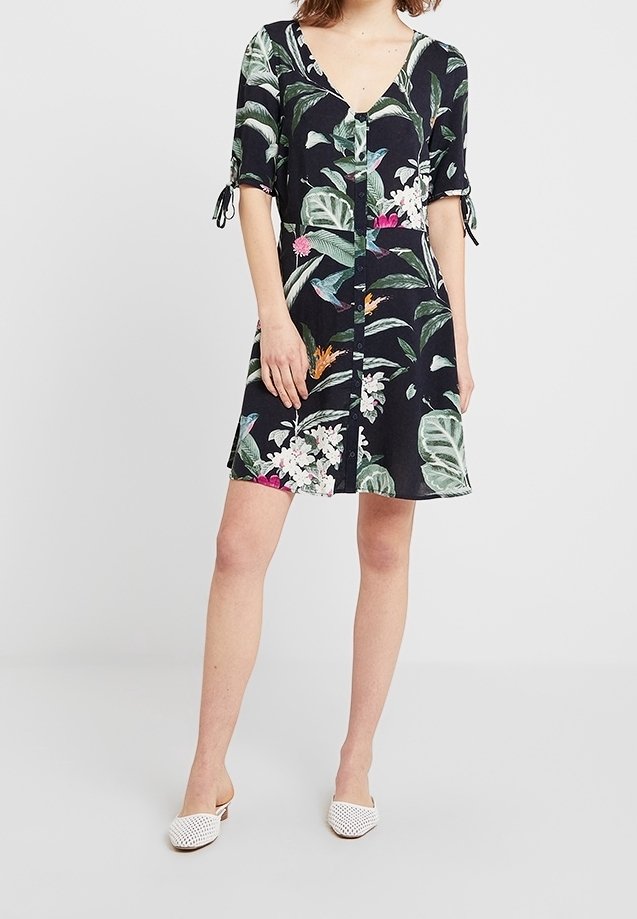 Femme portant une robe noire à manches courtes avec un imprimé de feuilles vertes et de fleurs, col en V, boutons à l'avant, et chaussures blanches sans lacets.