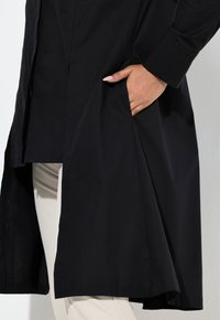 Osoba ubrana w długi czarny płaszcz z ręką w bocznej kieszeni i widocznymi jasnobeżowymi spodniami pod spodem.