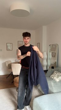 En marine genser holdes av en person, paret med svarte jeans og hvite sneakers. Lys, nøytral bakgrunn med minimal dekor.