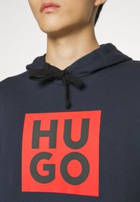 Gros plan d'une personne portant un sweat à capuche bleu marine avec des cordons noirs et un logo carré rouge affichant "HUGO" en lettres noires en gras.