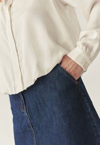 Biała bluzka z długimi rękawami i mankietami na guziki, zestawiona z jeansową spódnicą z widoczną kieszenią i kontrastującym przeszyciem.