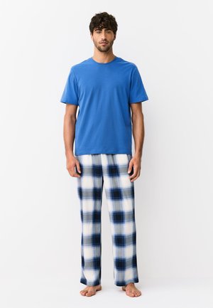 Homme debout pieds nus portant une chemise bleue unie à manches courtes et un pantalon de pyjama à carreaux noirs bleu-blanc sur fond blanc.