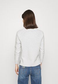 Hellgrauer Strickpullover mit einem gerippten Saum und Raglanärmeln, kombiniert mit blauen Jeans aus Denim, die ein Logopatch auf der Rückseite haben.