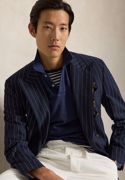 Tânăr purtând un blazer bleumarin cu dungi fine, un polo bleumarin cu cămașă în dungi dedesubt, pantaloni albi și un ceas de mână, așezat pe un fundal neutru.