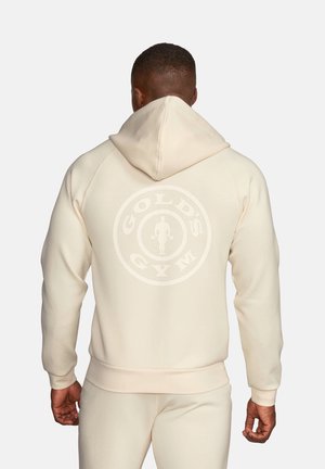 Mężczyzna w beżowej bluzie z kapturem i spodniach, widok od tyłu, logo Gold's Gym widoczne na plecach bluzy.