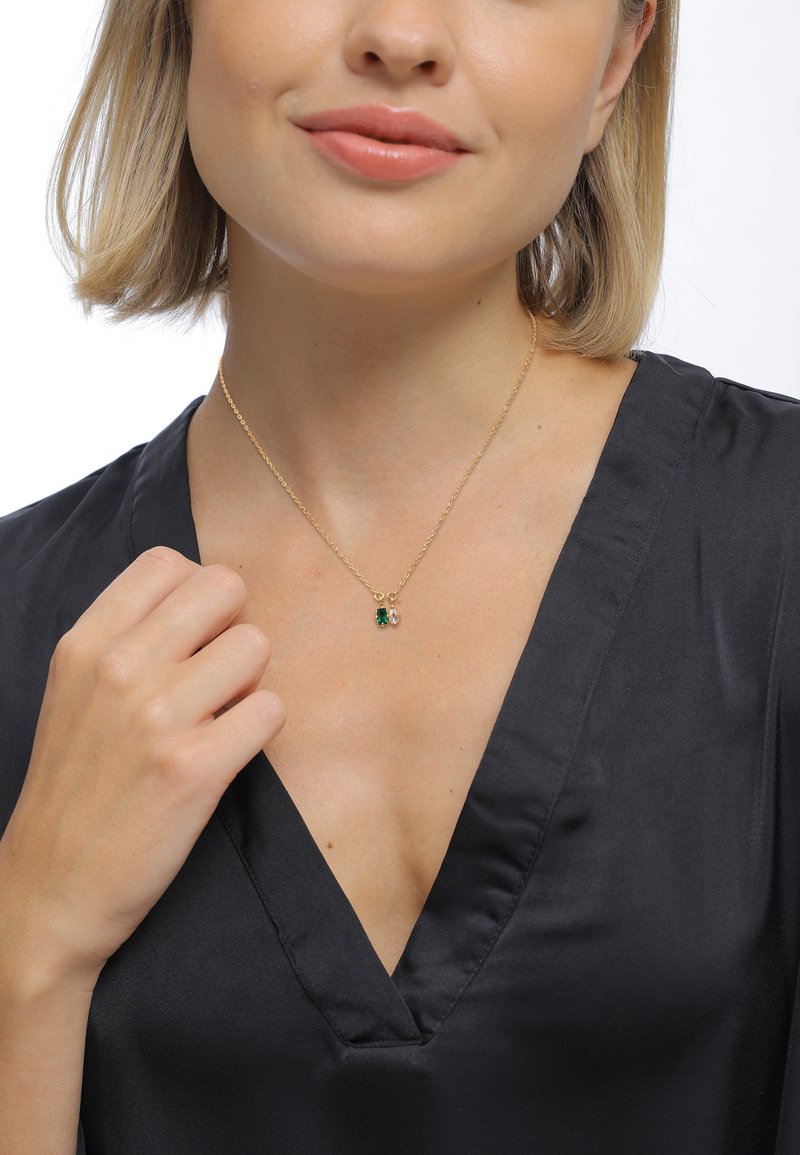 Guldkæde med vedhæng af grønne og klare krystaller. Modellen bærer en sort satin top, med hånden hvilende på kravebenet.