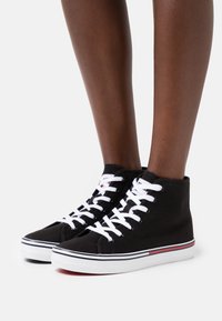 Czarne sneakersy za kostkę z płótna, z białymi sznurówkami, z białą podeszwą i czerwono-niebieskimi paskami na dole.