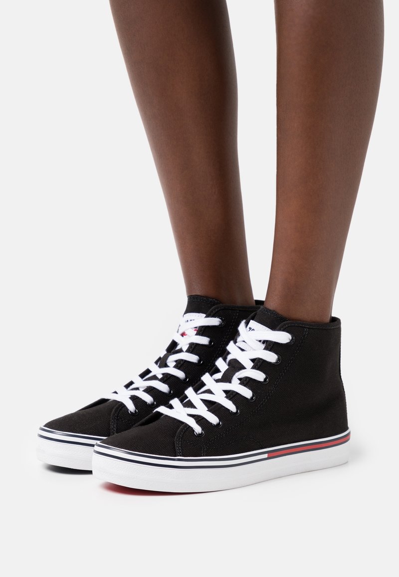 Czarne sneakersy za kostkę z płótna, z białymi sznurówkami, z białą podeszwą i czerwono-niebieskimi paskami na dole.