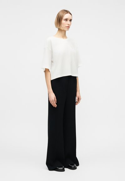 Γυναίκα με κοντά ξανθά μαλλιά που φοράει άσπρο μπλουζάκι με κοντά μανίκια, φαρδιά μαύρη παντελόνα και μαύρα παπούτσια, σε απλό λευκό φόντο.