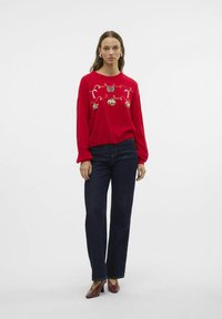 Maglione rosso con ricami decorativi, caratterizzato da ornamenti e bastoncini di zucchero, abbinato a jeans blu scuro e scarpe con tacco bordeaux.