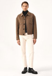 Chaqueta de lana marrón con botones, dos bolsillos en el pecho, llevada sobre un suéter de cuello alto negro, combinada con pantalones blancos y botines negros.