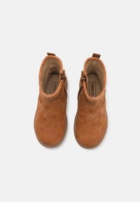 Botas de tobillo de cuero marrón con textura floral, cierre lateral y tira de agarre, mostradas desde una vista superior sobre un fondo blanco.