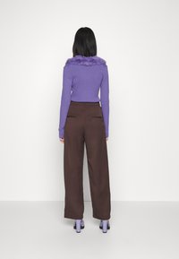 Femme portant un pull violet avec col en fourrure, pantalon large marron et chaussures à talons violettes, debout face à un mur blanc.