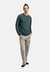 Camicia verde scuro a maniche lunghe con collo a coste, abbinata a pantaloni beige chiaro con tasche laterali e sneakers nere con suole bianche.