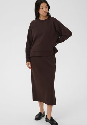 Femme portant un sweat-shirt marron foncé à manches longues et une jupe assortie longueur cheville avec des chaussures noires à enfiler, debout avec une main sur la hanche.