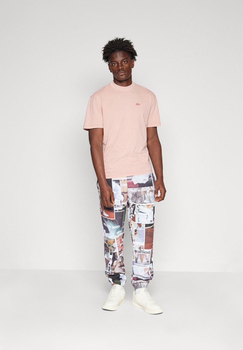 T-shirt en coton rose clair à manches courtes avec un logo discret, assortie à un pantalon multicolore à motifs et des baskets blanches.