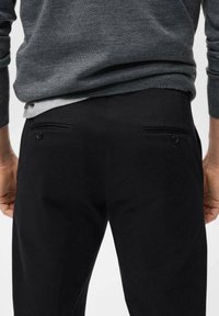 Pantalones negros con una tela suave y un corte ajustado, bolsillos traseros con cierres de botón y una cinturilla visible debajo de un suéter gris.