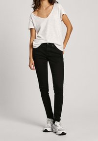 Kvinna som bär en lös vit T-shirt med V-ringning, svarta skinny jeans och vita sneakers, stående med ena handen i fickan.