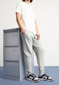 White cotton t-shirt with short sleeves, paired with light grey sweatpants and black and white Nike sneakers, standing beside a grey filing cabinet.