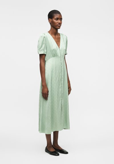 Γυναίκα με ανοιχτοπράσινο μίντι φόρεμα με φουσκωτά μανίκια και κουμπιά μπροστά, συνδυασμένο με μαύρα μπαλαρίνες, στέκεται μπροστά σε μονόχρωμο φόντο.