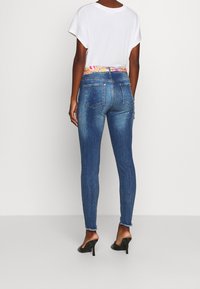 Úzké modré džíny se vzorovaným pasem a dekorativními akcenty. Nosené s volným bílým tričkem a černými lodičkami.