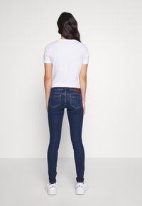 Jeans ajustados de mezclilla oscura con una textura suave, bolsillos traseros detallados y una etiqueta de cuero marrón en la cintura, combinados con una camiseta blanca.